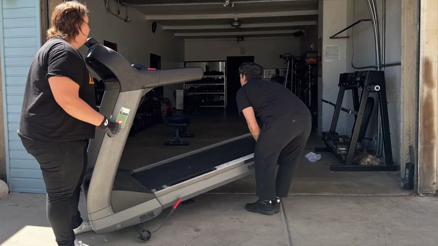 Two workers wheeling an old treadmill out of a home gym on a dolly