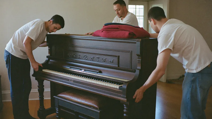 Three workers rolling an old upright piano on a dolly toward the front door